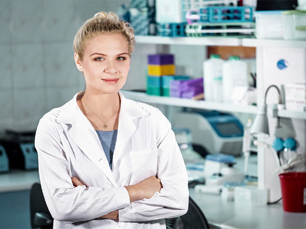 Kobieta w białym fartuchu, ze skrzyżowanymi ramionami, na tle regałów ze sprzętem laboratoryjnym