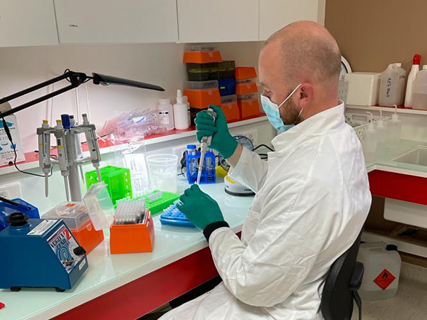 Mężczyzna w białym fartuchu siedzi przy stole laboratoryjnym. Wstrzykuje pipetą płyn do małej probówki