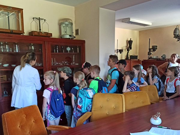 Kobieta w białym fartuchu stoi wśród dzieci w wieku szkolnym. Wszyscy patrzą się na zawartość przeszklonej gabloty