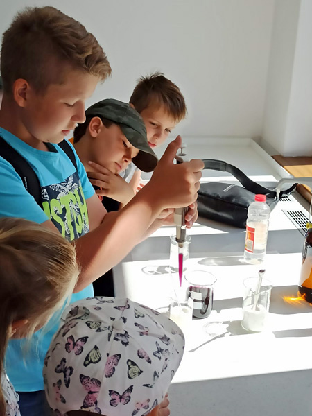 Chłopiec odmierza pipetą porcję fioletowego płynu. Koledzy i koleżanki obserwują, co robi