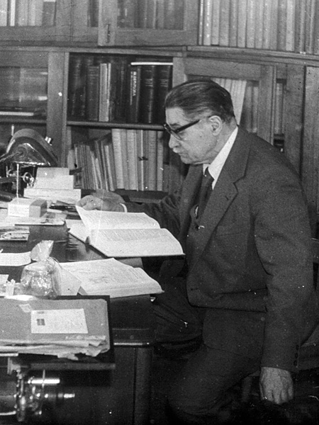 Czarnobiała fotografia starszego mężczyzny w okularach czytającego przy biurku grubą książkę. W tle regały biblioteczne