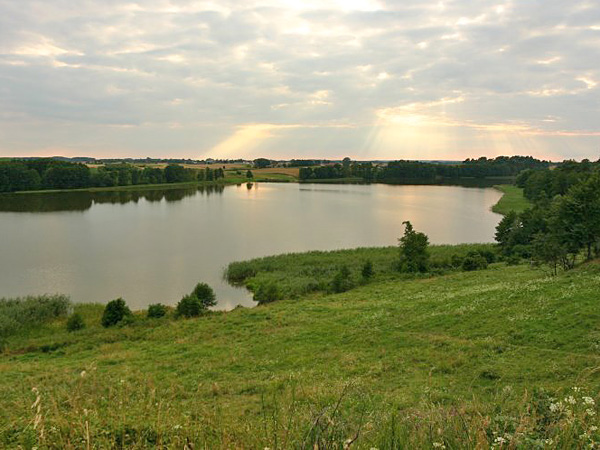 A view from a grassy hillside over a lake in an idyllic, agricultural landscape. Sunlight shines through the clouds