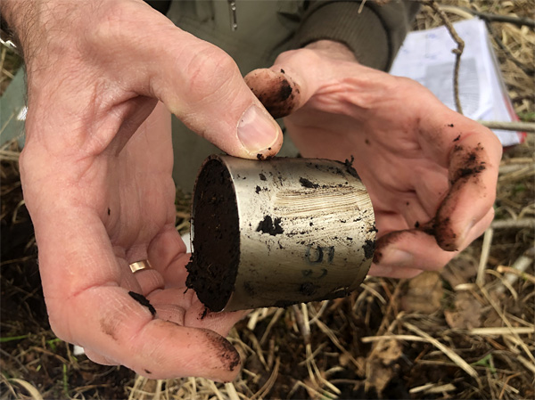 Ubrudzone dłonie męskie trzymają niewielki metalowy cylinder wypełniony i umazany z zewnątrz czarną glebą