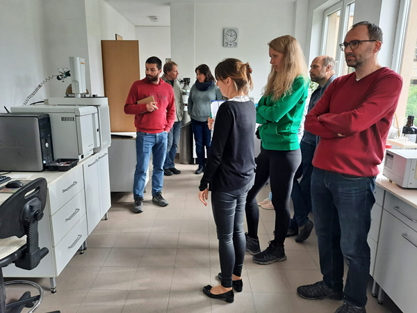 Grupa osób zwiedza jasne pomieszczenie laboratoryjne. Przy ścianach białe szafki i aparatura naukowa