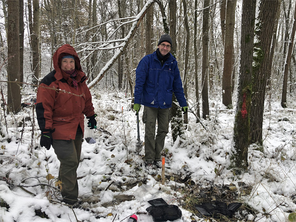 Kobieta i mężczyzna stoją w zimowych kurtkach w ośnieżonym lesie. U ich stóp odgarnięty śnieg i czarne kwadratowe woreczki