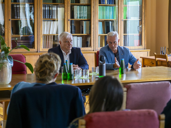 Dwóch mężczyzn siedzi za dużym drewnianym stołem. Jeden gestykulując, mówi w kierunku publiczności