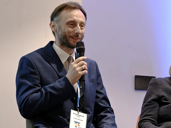 Mężczyzna w garniturze, pokazany od pasa w górę, mówi przez mikrofon, uśmiechając się do publiczności