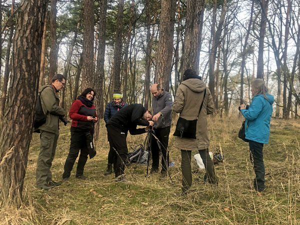 Grupa ludzi w strojach terenowych stoi pośród sosen wokół urządzenia zamontowanego na statywie